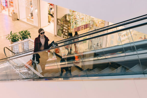 Eine Frau mit Sonnenbrille und Einkaufstaschen fährt mit der Rolltreppe in einem Einkaufszentrum, im Hintergrund sind verschiedene Geschäfte zu sehen.