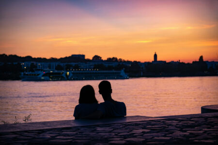 Ein Paar sitzt am Ufer des Rheins und schaut bei Sonnenuntergang auf das Wasser, im Hintergrund ist ein beleuchtetes Schiff und die Skyline von Duisburg zu sehen.