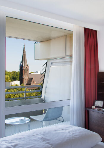 Hotelzimmer im Hotel Rheingarten in Duisburg mit einem Balkon und Blick auf eine Kirche, ausgestattet mit modernen Möbeln und einem Schreibtisch.