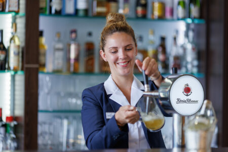 Barkeeperin im Hotel Rheingarten in Duisburg zapft ein Bier und lächelt, im Hintergrund Flaschenregal mit verschiedenen Getränken.