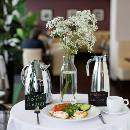 Frühstückstisch im Hotel Rheingarten in Duisburg mit einer Vase mit Blumen, Kaffeekannen und einem Teller mit Spiegelei und Gemüse.