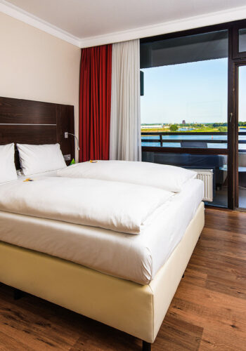 Modern eingerichtetes Hotelzimmer im Hotel Rheingarten in Duisburg mit einem Doppelbett und Balkon mit Blick auf den Rhein.